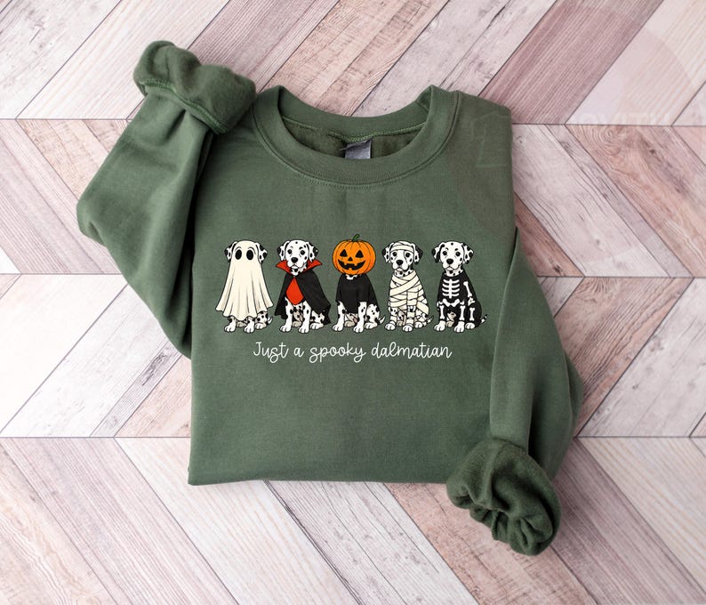May include: An olive green crewneck sweatshirt featuring a graphic of five Dalmatian dogs in various Halloween costumes, including a ghost, vampire, pumpkin head, mummy, and skeleton. The text below the dogs reads, Just a spooky dalmatian.