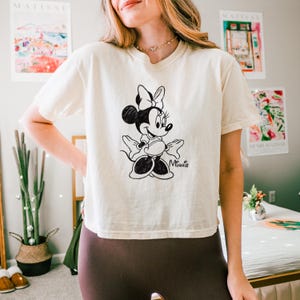 May include: Cream-colored t-shirt featuring a black and white illustration of Minnie Mouse. The graphic shows Minnie with her signature bow and a playful pose. The word "Minnie" is written in script below the character.