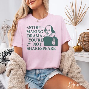 May include: A light pink short-sleeve t-shirt featuring a dark green graphic of William Shakespeare with a hand raised in a stop gesture. The text reads: STOP MAKING DRAMA YOU'RE NOT SHAKESPEARE, accented with small four-pointed star icons.