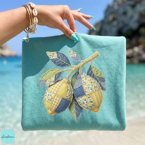 May include: A turquoise zippered pouch featuring a lemon branch applique. The lemons are patchwork with yellow floral and gingham patterns, and denim blue accents. The leaves are green and blue. The pouch is held by a hand with turquoise nails.