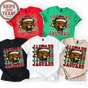 May include: A collection of t-shirts in various colors, including red, green, black, white, and tan. Each shirt features a jaguar head wearing a Santa hat, with the text "Tis the season" and "Jaguars" in a festive design.
