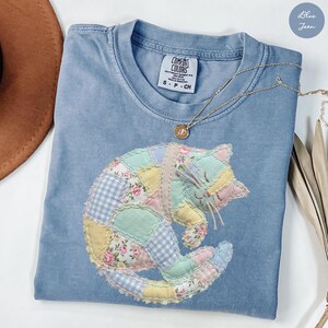May include: A blue Comfort Colors t-shirt with a patchwork cat design. The cat is made of various pastel fabrics, including floral and gingham patterns. A gold necklace with a pendant rests on the shirt. The shirt is labeled "Blue Jean".