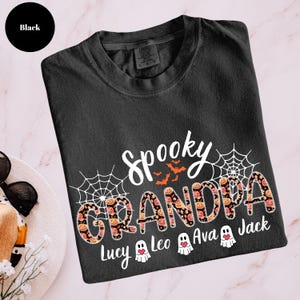 Może przedstawiać: Czarna koszulka z białym napisem "Spooky Grandpa", z pomarańczowymi nietoperzami i pajęczynami. Słowo "Grandpa" jest ozdobione dyniami i kwiatami. Poniżej znajdują się imiona Lucy, Leo, Ava i Jack.