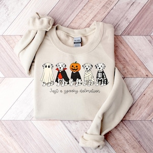 May include: A cream-colored crewneck sweatshirt featuring a graphic of five Dalmatian dogs in various Halloween costumes, including a ghost, vampire, pumpkin, mummy, and skeleton, with the text "Just a spooky dalmatian" printed below.