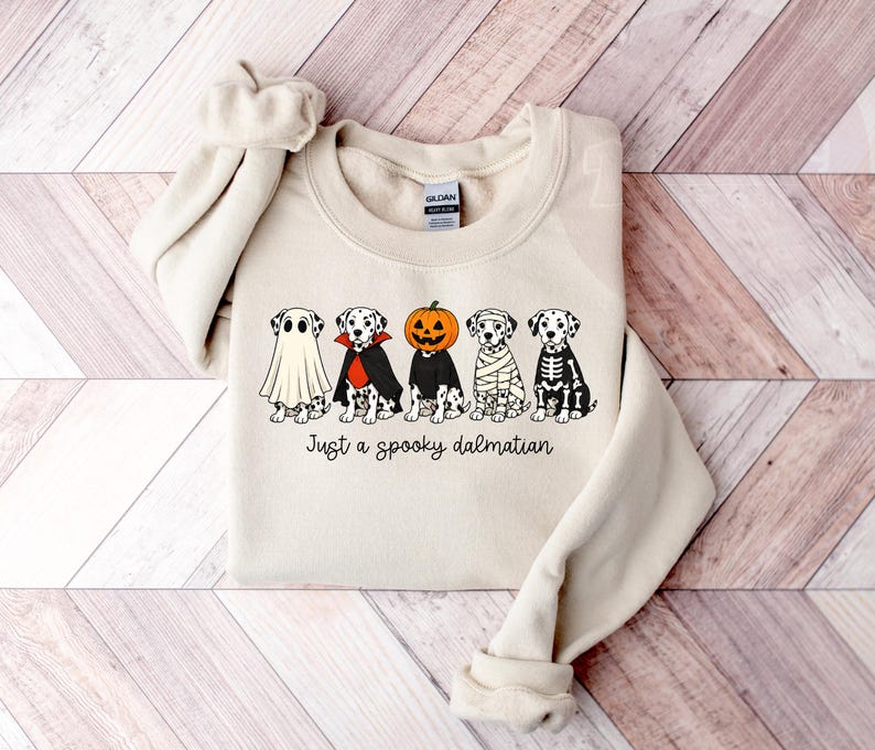 May include: A cream-colored crewneck sweatshirt featuring a graphic of five Dalmatian dogs in various Halloween costumes, including a ghost, vampire, pumpkin, mummy, and skeleton, with the text "Just a spooky dalmatian" printed below.