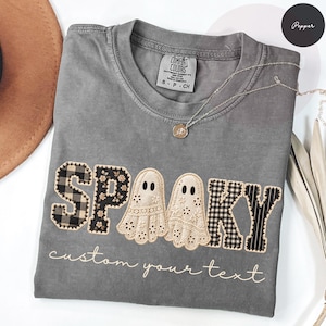 May include: A pepper-coloured crewneck t-shirt featuring an embroidered design with the word SPOOKY, where the letters are replaced by two cream-coloured lace ghosts. The letters S, P, and KY feature black and white plaid and floral patterns.