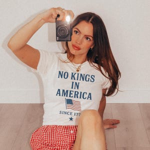 May include: White t-shirt with the text "NO KINGS IN AMERICA" and an American flag graphic. The shirt is paired with red and white checkered shorts. A black camera is held up with a flash.