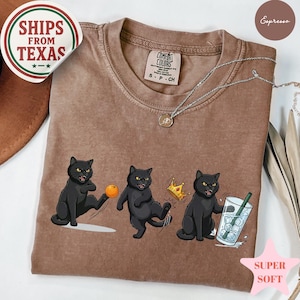Puede incluir: Camiseta color espresso con un gr&aacute;fico de tres gatos negros en varias poses. Un gato patea una pelota, otro lleva una corona y el &uacute;ltimo sostiene una bebida. La camiseta tiene una etiqueta Comfort Colors y las palabras "SUPER SOFT".