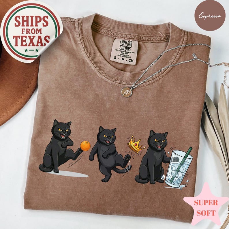 Puede incluir: Camiseta color espresso con un gr&aacute;fico de tres gatos negros en varias poses. Un gato patea una pelota, otro lleva una corona y el &uacute;ltimo sostiene una bebida. La camiseta tiene una etiqueta Comfort Colors y las palabras "SUPER SOFT".