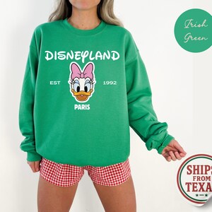 Puede incluir: Una sudadera verde de Disneyland con un gr&aacute;fico de Daisy Duck. La sudadera tiene el texto "Disneyland" encima del gr&aacute;fico, con "Est 1992 Paris" debajo. La sudadera se combina con unos shorts a cuadros rojos y blancos.
