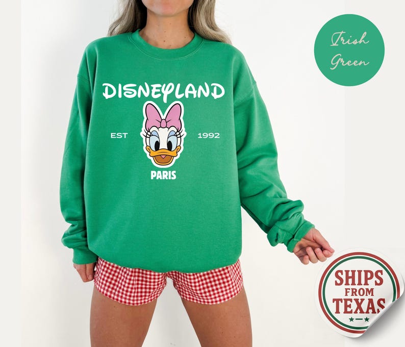 Puede incluir: Una sudadera verde de Disneyland con un gr&aacute;fico de Daisy Duck. La sudadera tiene el texto "Disneyland" encima del gr&aacute;fico, con "Est 1992 Paris" debajo. La sudadera se combina con unos shorts a cuadros rojos y blancos.