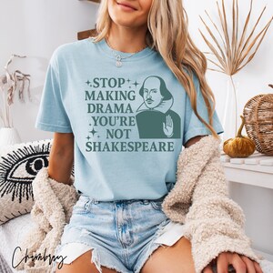 May include: A light blue short-sleeve t-shirt featuring a dark green graphic of William Shakespeare with a raised hand and the text "STOP MAKING DRAMA YOU'RE NOT SHAKESPEARE" surrounded by small star icons printed on the front of the garment.