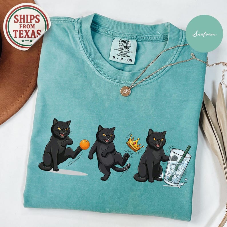 Puede incluir: Camiseta verde mar con tres ilustraciones de gatos negros. Un gato patea una pelota naranja, otro lleva una corona y el tercero sostiene un vaso de agua con hielo. Tambi&eacute;n se ven un collar dorado y una pegatina "Ships from Texas".