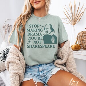 May include: A sage green short-sleeve t-shirt featuring a dark green graphic of William Shakespeare with a raised hand. The text reads: STOP MAKING DRAMA YOU'RE NOT SHAKESPEARE, accented with small four-pointed star icons on the left side of the print.