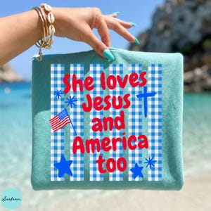 May include: A teal-colored zippered pouch with the text "She loves Jesus and America too" in red and blue. The design includes a blue and white gingham pattern, a cross, an American flag, and blue stars.