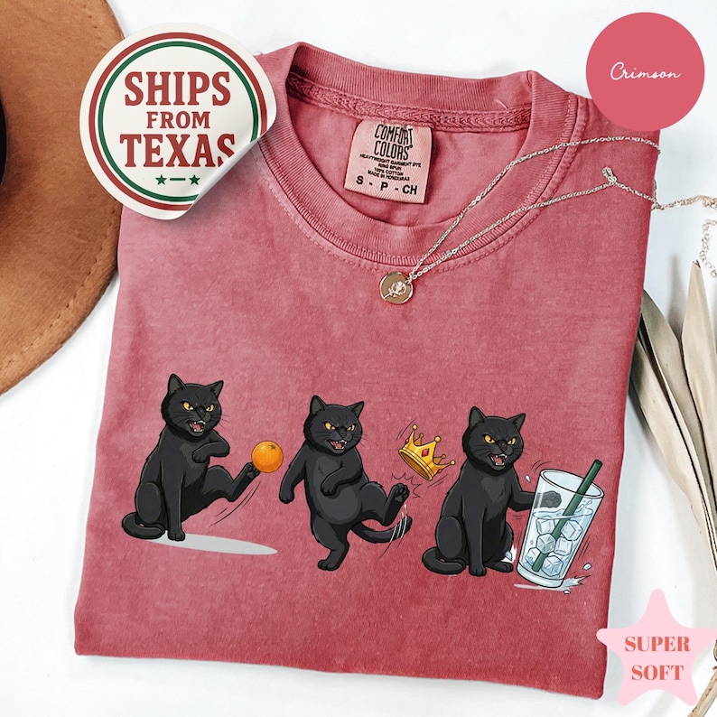 Puede incluir: Camiseta de color carmes&iacute; con una caricatura de tres gatos negros. Uno patea una pelota naranja, otro lleva una corona y el tercero sostiene una bebida. La camiseta tiene una etiqueta Comfort Colors y una pegatina Ships From Texas.