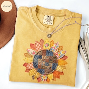 Puede incluir: Camiseta Comfort Colors de color mostaza con un gran diseño de girasol aplicado. El girasol presenta varias telas estampadas en tonos de amarillo, naranja y marrón. Un collar de plata con un pequeño colgante descansa sobre la camisa.