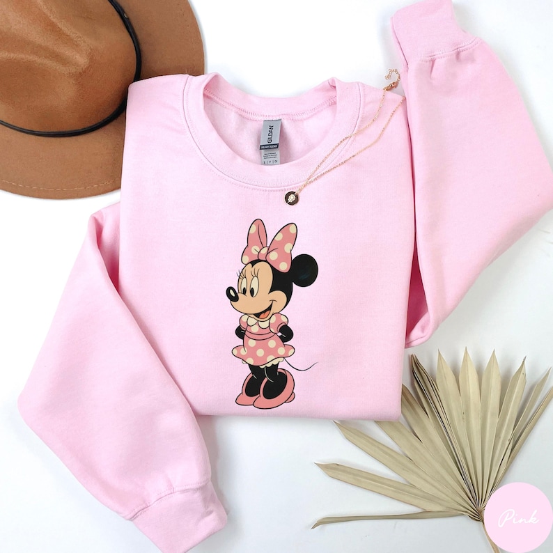 May include: A light pink sweatshirt featuring a Minnie Mouse graphic. The cartoon character is wearing a pink dress with white polka dots and a matching bow. A gold necklace and a brown hat are also visible.