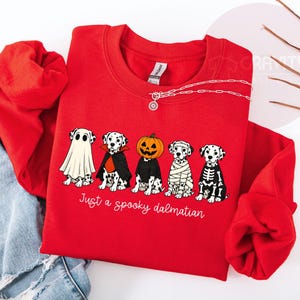 May include: A bright red crewneck sweatshirt featuring a graphic of five Dalmatian dogs in various Halloween costumes, including a ghost, vampire, pumpkin head, mummy, and skeleton. The text below the dogs reads Just a spooky dalmatian.