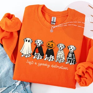 May include: An orange crewneck sweatshirt featuring a graphic of five Dalmatian dogs dressed in Halloween costumes: a ghost, a vampire, a pumpkin, a mummy, and a skeleton. Below the dogs, the text reads, "Just a spooky dalmatian" in white script.