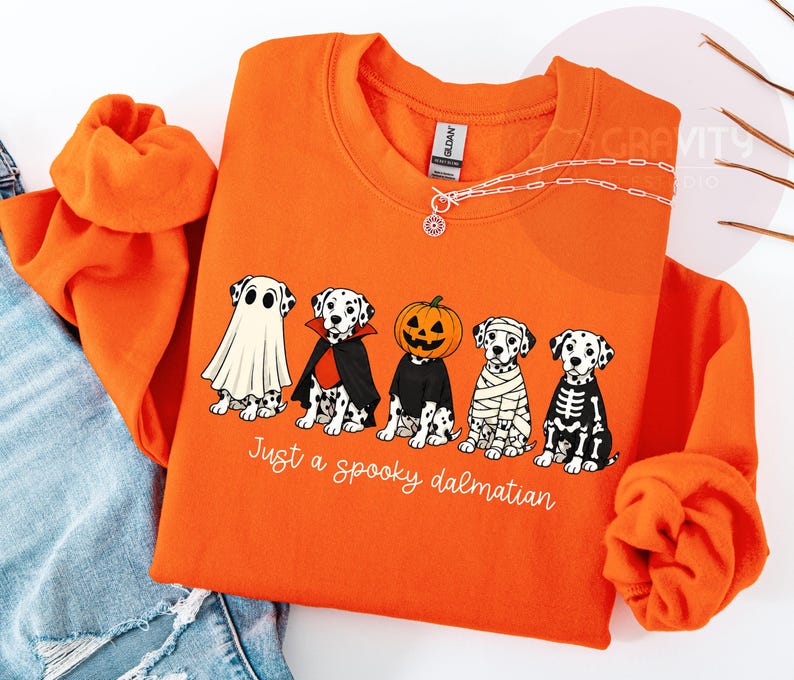 May include: An orange crewneck sweatshirt featuring a graphic of five Dalmatian dogs dressed in Halloween costumes: a ghost, a vampire, a pumpkin, a mummy, and a skeleton. Below the dogs, the text reads, "Just a spooky dalmatian" in white script.