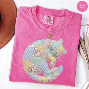 May include: A pink Comfort Colors t-shirt featuring a patchwork cat design. The cat is made of various pastel fabrics, including floral and gingham patterns. A gold necklace with a pendant rests on the shirt. The shirt is size S-P-CH.