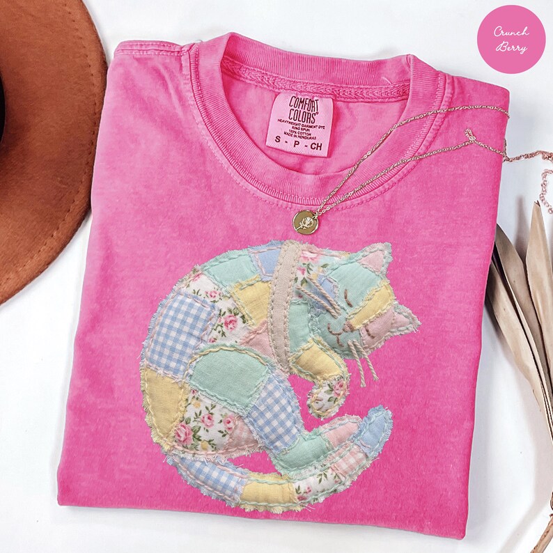 May include: A pink Comfort Colors t-shirt featuring a patchwork cat design. The cat is made of various pastel fabrics, including floral and gingham patterns. A gold necklace with a pendant rests on the shirt. The shirt is size S-P-CH.