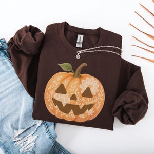 May include: A brown sweatshirt featuring a large, orange pumpkin design with a carved face and floral accents. The pumpkin has a green stem and is set against a dark background. A silver necklace is draped across the sweatshirt.