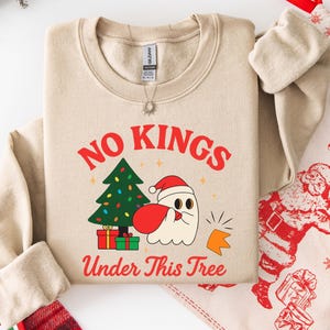 Può includere: Felpa beige con la scritta rossa "NO KINGS UNDER THIS TREE". La grafica include un albero di Natale, un fantasma con cappello da Babbo Natale e regali. È visibile una collana d'argento.