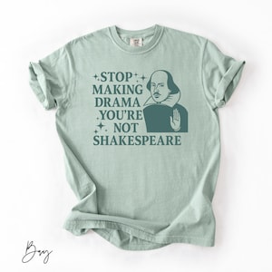May include: A light sage green short-sleeve t-shirt featuring a dark green graphic of William Shakespeare with his hand raised. The text reads: STOP MAKING DRAMA YOU'RE NOT SHAKESPEARE, accented by small four-pointed star icons on the left side.