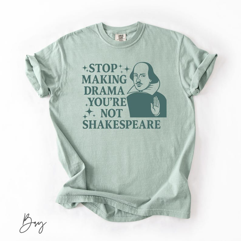 May include: A light sage green short-sleeve t-shirt featuring a dark green graphic of William Shakespeare with his hand raised. The text reads: STOP MAKING DRAMA YOU'RE NOT SHAKESPEARE, accented by small four-pointed star icons on the left side.
