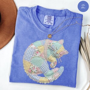 May include: A blue Comfort Colors t-shirt featuring a patchwork cat design in pastel colors. The cat is curled up, with a mix of floral, gingham, and solid fabric patches. A silver necklace with a pendant rests on the shirt.