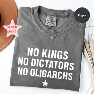 Puede incluir: Una camiseta gris con el texto blanco "NO KINGS NO DICTATORS NO OLIGARCHS". La camiseta tiene cuello redondo y mangas cortas. Un collar con colgante dorado está drapeado sobre la camiseta. La camiseta está etiquetada como "Pepper".