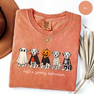 Może przedstawiać: Koszulka z okrągłym dekoltem w kolorze terakoty z grafiką przedstawiającą pięć dalmatyńczyków w kostiumach na Halloween, w tym ducha, wampira, dynię, mumię i szkielet. Poniżej znajduje się biały napis Just a spooky dalmatian.