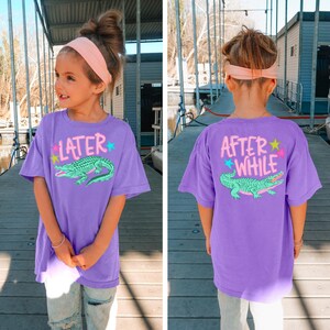 May include: A purple t-shirt with the text "LATER" and a green alligator graphic on the front. The back of the shirt has the text "AFTER WHILE" and a matching alligator design. The shirt is worn by a young person.