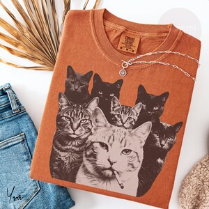 May include: A rust-colored t-shirt featuring a graphic print of seven cats, including one in the foreground with a cigarette in its mouth. The shirt has a crew neckline and a Comfort Colors brand label on the inside collar area.