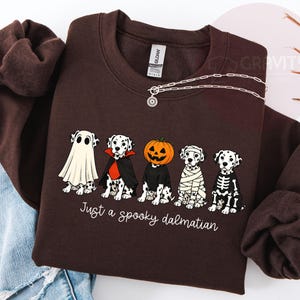 May include: A dark brown crewneck sweatshirt featuring a graphic of five Dalmatian dogs in Halloween costumes, including a ghost, a vampire, a pumpkin head, a mummy, and a skeleton. The text below the dogs reads, Just a spooky dalmatian.