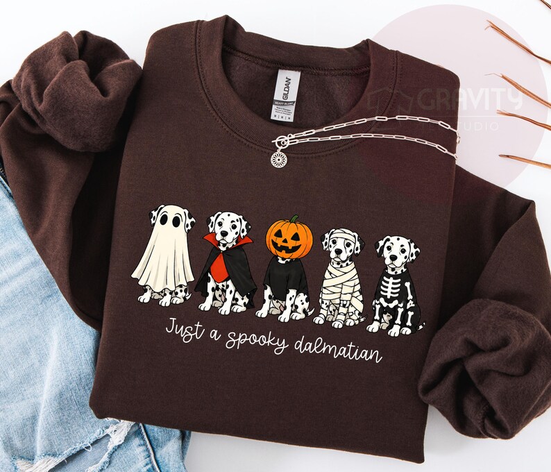 May include: A dark brown crewneck sweatshirt featuring a graphic of five Dalmatian dogs in Halloween costumes, including a ghost, a vampire, a pumpkin head, a mummy, and a skeleton. The text below the dogs reads, Just a spooky dalmatian.