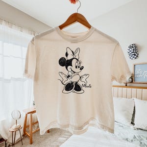 May include: Cream-colored t-shirt featuring a black and white illustration of Minnie Mouse. The graphic shows Minnie with her signature bow and gloves. The word "Minnie" is written in cursive below the character. The shirt is hanging on a wooden hanger.