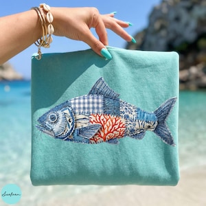 Puede incluir: Una bolsa de cremallera turquesa con un diseño de pez aplicado. El pez está compuesto por varias telas azules estampadas, incluyendo cuadros y estampados florales, con acentos de coral rojo. La bolsa se muestra sobre un fondo de playa.