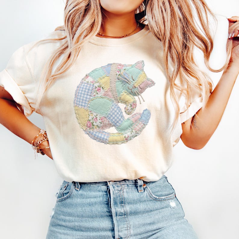 May include: Cream-colored t-shirt featuring a patchwork cat design. The cat is composed of various pastel-colored fabrics, including floral prints, gingham, and solid colors. The cat is curled up, with its eyes closed, giving a whimsical and cozy feel.