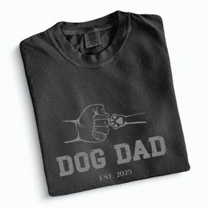 May include: Dark grey t-shirt featuring a graphic of a fist bump with a dog's paw. The text "DOG DAD" is printed below the graphic, with "EST. 2025" underneath. The shirt is folded and has a tag in the upper left corner.