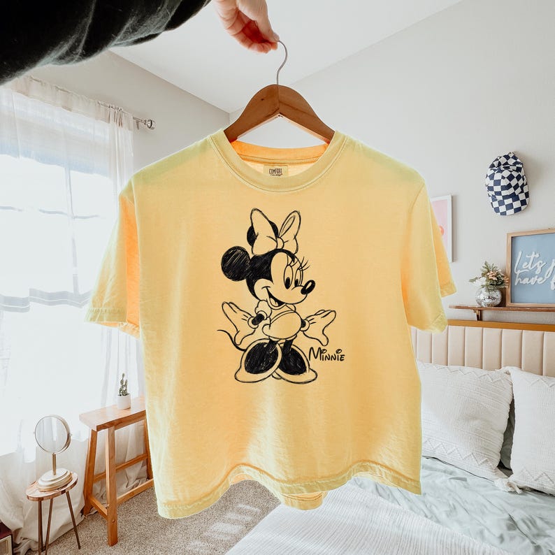 May include: Yellow t-shirt featuring a black and white illustration of Minnie Mouse. The graphic shows Minnie Mouse with her signature bow and polka dot skirt. The word "Minnie" is printed below the character.