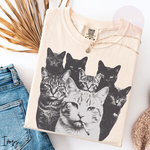 May include: A light beige t-shirt featuring a black and white graphic print of seven cats, including one cat in the foreground holding a cigarette in its mouth. The shirt has a Comfort Colors brand label at the inner neckline and a crew neck collar.