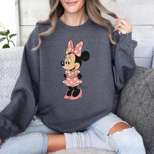 May include: A charcoal gray sweatshirt featuring a Minnie Mouse graphic. Minnie is depicted in a pink polka dot dress and bow. The sweatshirt has long sleeves and a crew neck.