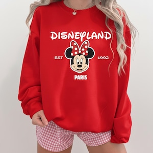 Puede incluir: Una sudadera roja de Disneyland Par&iacute;s con Minnie Mouse y un lazo de lunares. La sudadera muestra el texto "Disneyland", "EST 1992" y "PARIS". Un dise&ntilde;o cl&aacute;sico para los fans de Disney.