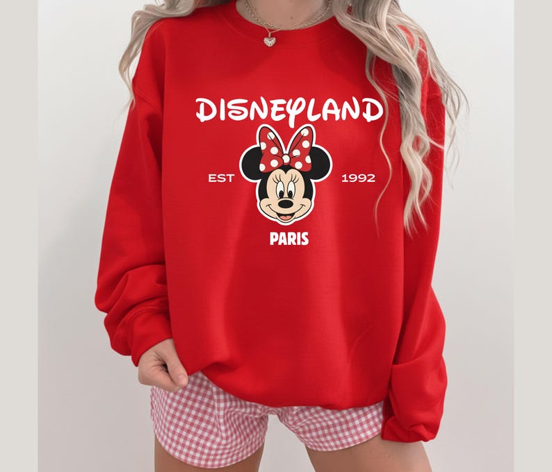 Puede incluir: Una sudadera roja de Disneyland Par&iacute;s con Minnie Mouse y un lazo de lunares. La sudadera muestra el texto "Disneyland", "EST 1992" y "PARIS". Un dise&ntilde;o cl&aacute;sico para los fans de Disney.
