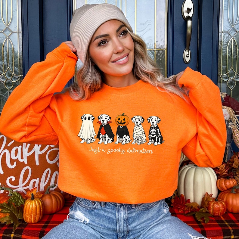 May include: A bright orange crewneck sweatshirt featuring a graphic of five Dalmatian dogs in various Halloween costumes, including a ghost, vampire, pumpkin, mummy, and skeleton, with the text Just a spooky dalmatian printed below.