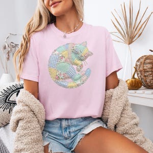 May include: A light pink t-shirt featuring a patchwork cat design. The cat is composed of various pastel-colored fabrics, including floral and gingham patterns. The cat is curled up, with its eyes closed, giving a cozy and whimsical feel.
