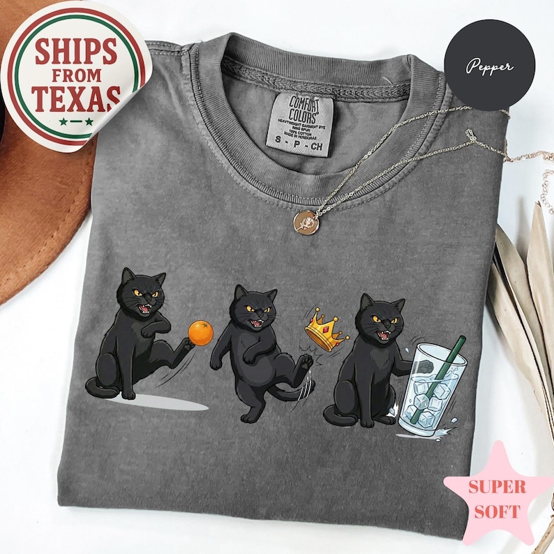Puede incluir: Camiseta gris Comfort Colors con un gr&aacute;fico de tres gatos negros. Un gato patea una pelota naranja, otro lleva una corona y el &uacute;ltimo sostiene una bebida con hielo. Un collar plateado est&aacute; drapeado sobre la camiseta.